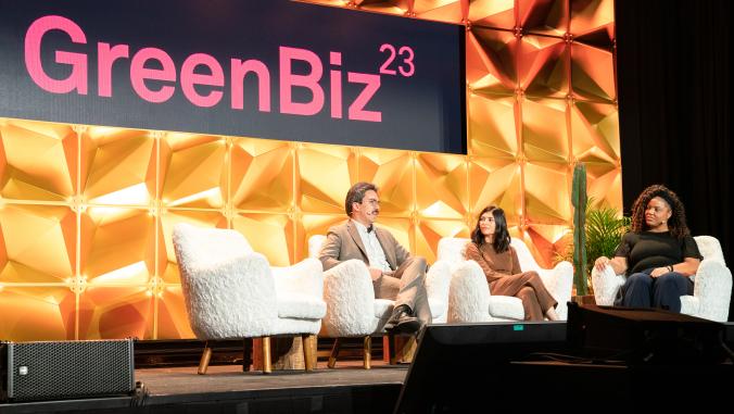 A picture of two women and one man sitting in white chairs on a stage. Behind them is a gold wall with the words "GreenBiz23" projected in pink.